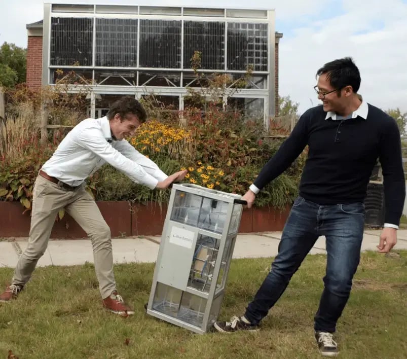 Two men outdoors lifting a prototype of Aquabattery's sustainable energy storage system, smiling as they tilt it upright in front of a building with large windows and garden plants.