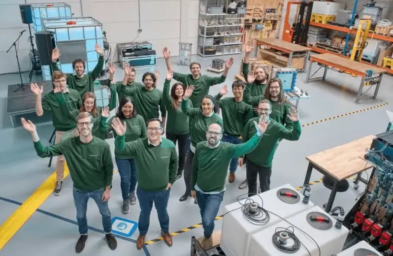 The Aquabattery team stand and wave at the camera from an industrial research environment.