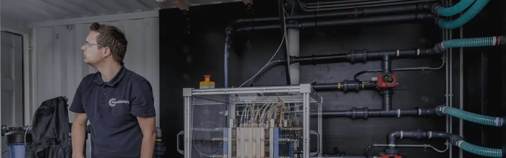 A man stands next to Aquabattery's flow battery system, wearing a navy Aquabattery polo shirt and looking off-camera to his right.