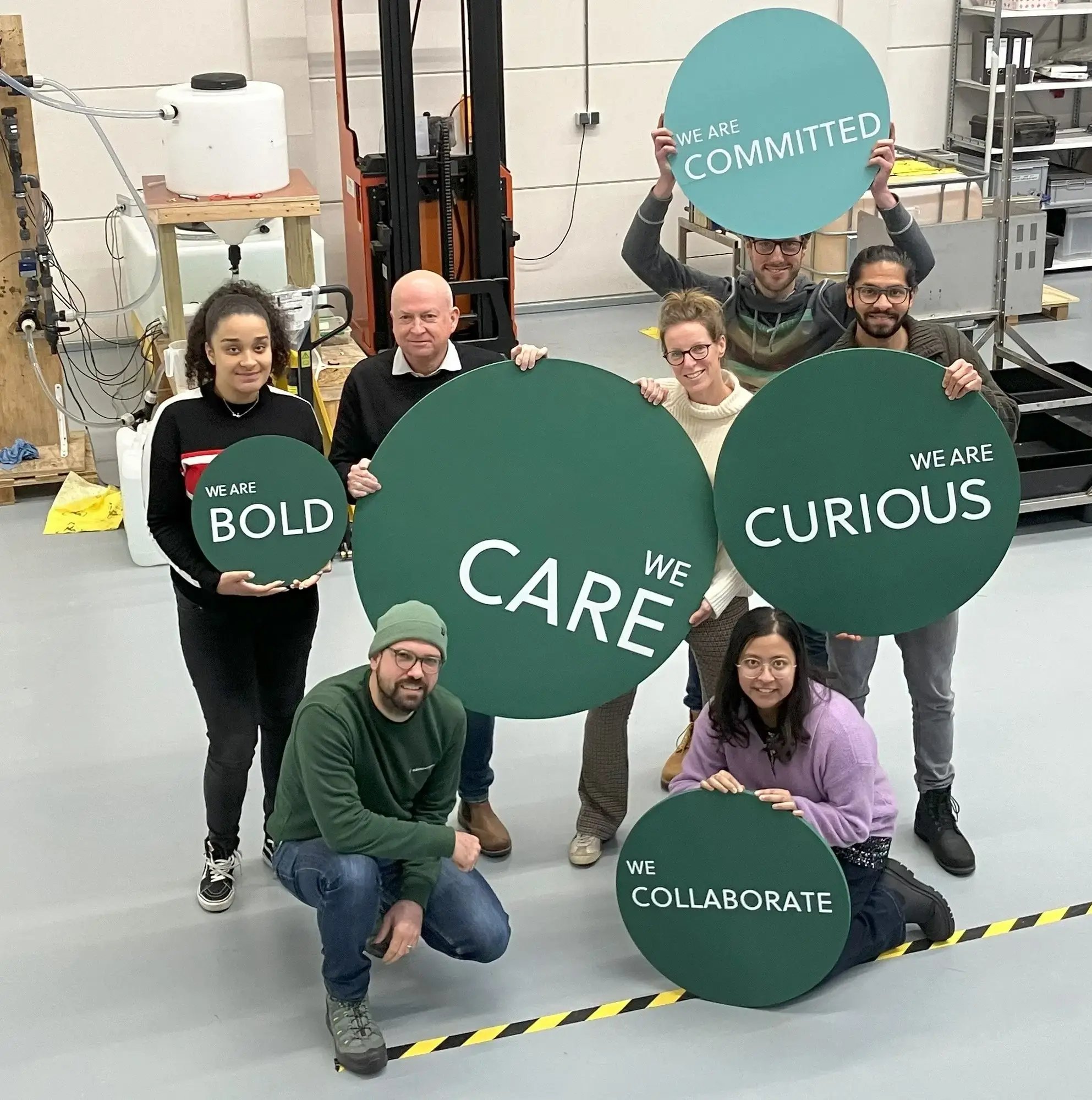 Members of the Aquabattery team hold up signs with company values written on them. These values are: WE ARE COMMITTED, WE ARE BOLD, WE ARE CURIOUS, WE CARE and WE COLLABORATE.