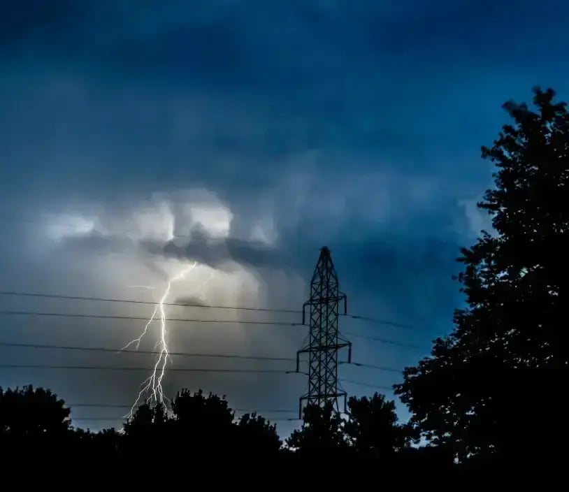 A bolt of lightning close to an electricity pylon against a stormy night sky, with trees in the foreground.
